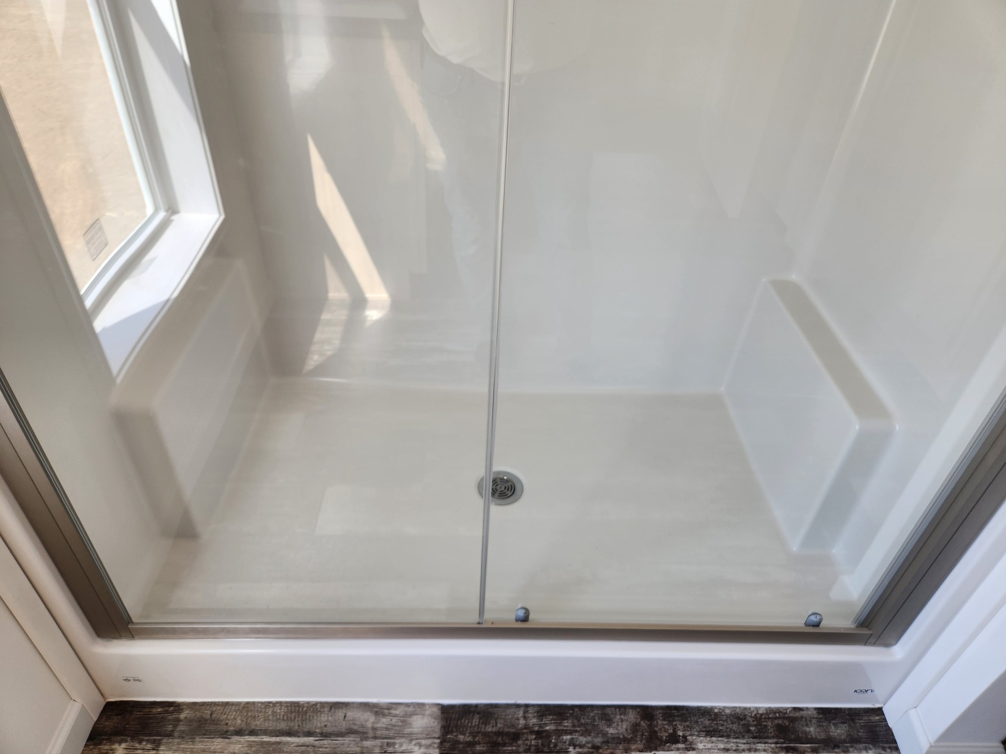 A clean, white shower stall with a glass door, set in a bathroom. It has a small step-in ledge, a drain in the center, and a window on the left side.