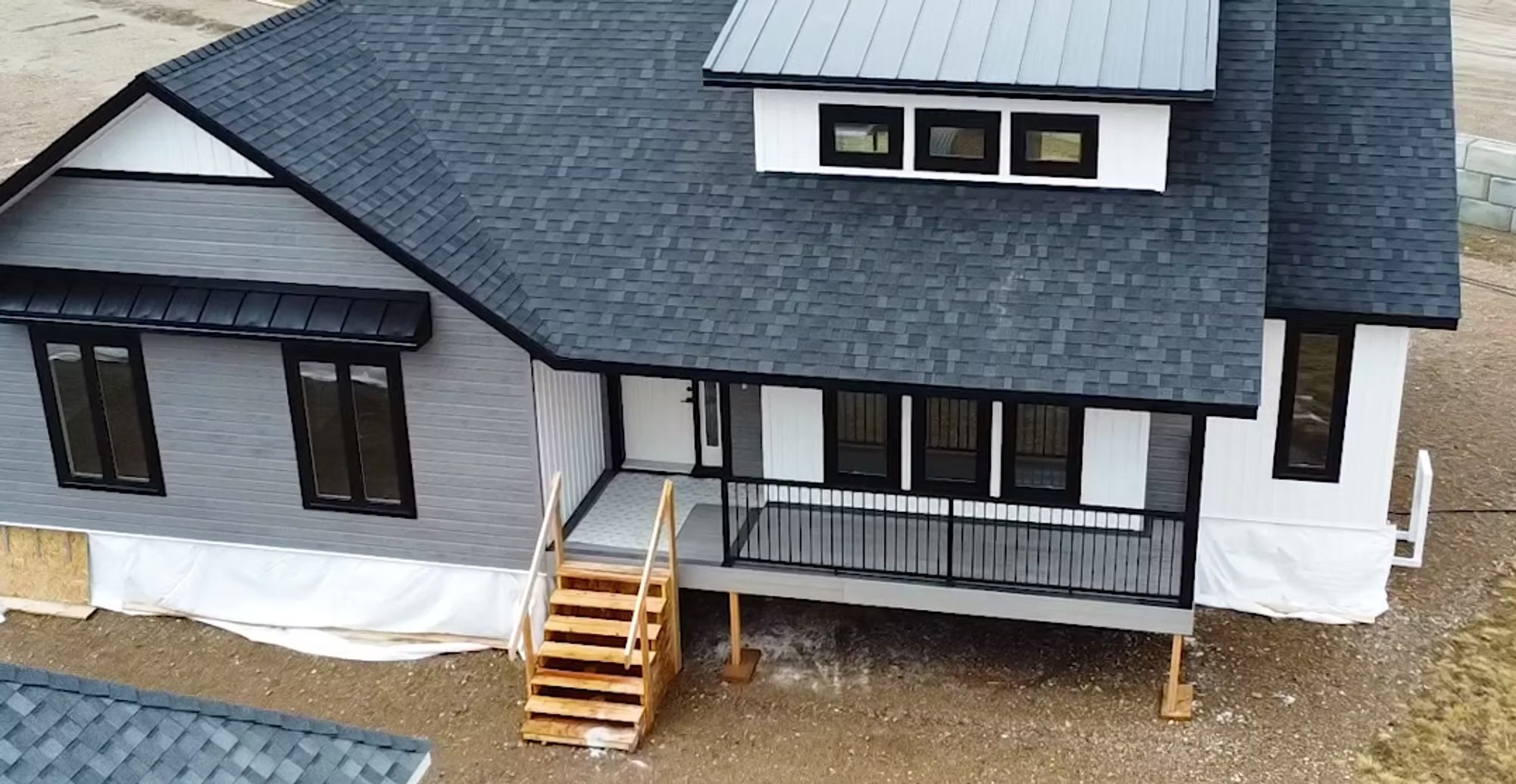 A modern house with dark shingles, white and gray siding, and black window frames. It features a covered porch and wooden steps, conveying a minimalist style.