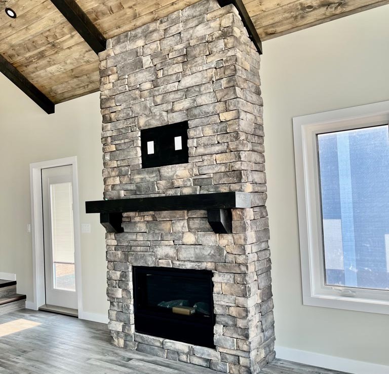 Rustic grey stone fireplace with a black mantel in a cozy living room. Vaulted wooden ceiling and large windows enhance the warm, inviting space.