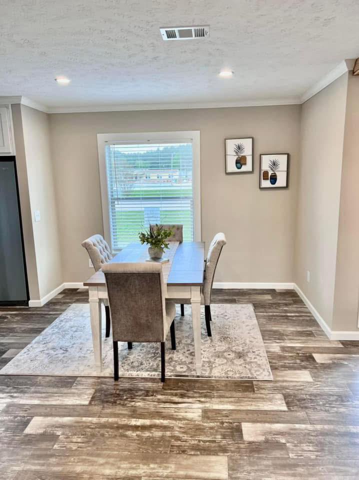 A modern dining area with a wooden table and four chairs on a patterned rug, under bright lighting. A window view and two plant-themed artworks add charm.