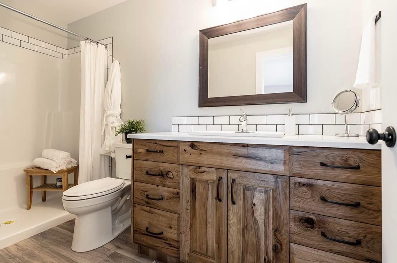 Rustic bathroom with wood vanity, white countertop, large mirror, and tiled backsplash. Shower with curtain and wooden stool; clean, cozy ambiance.