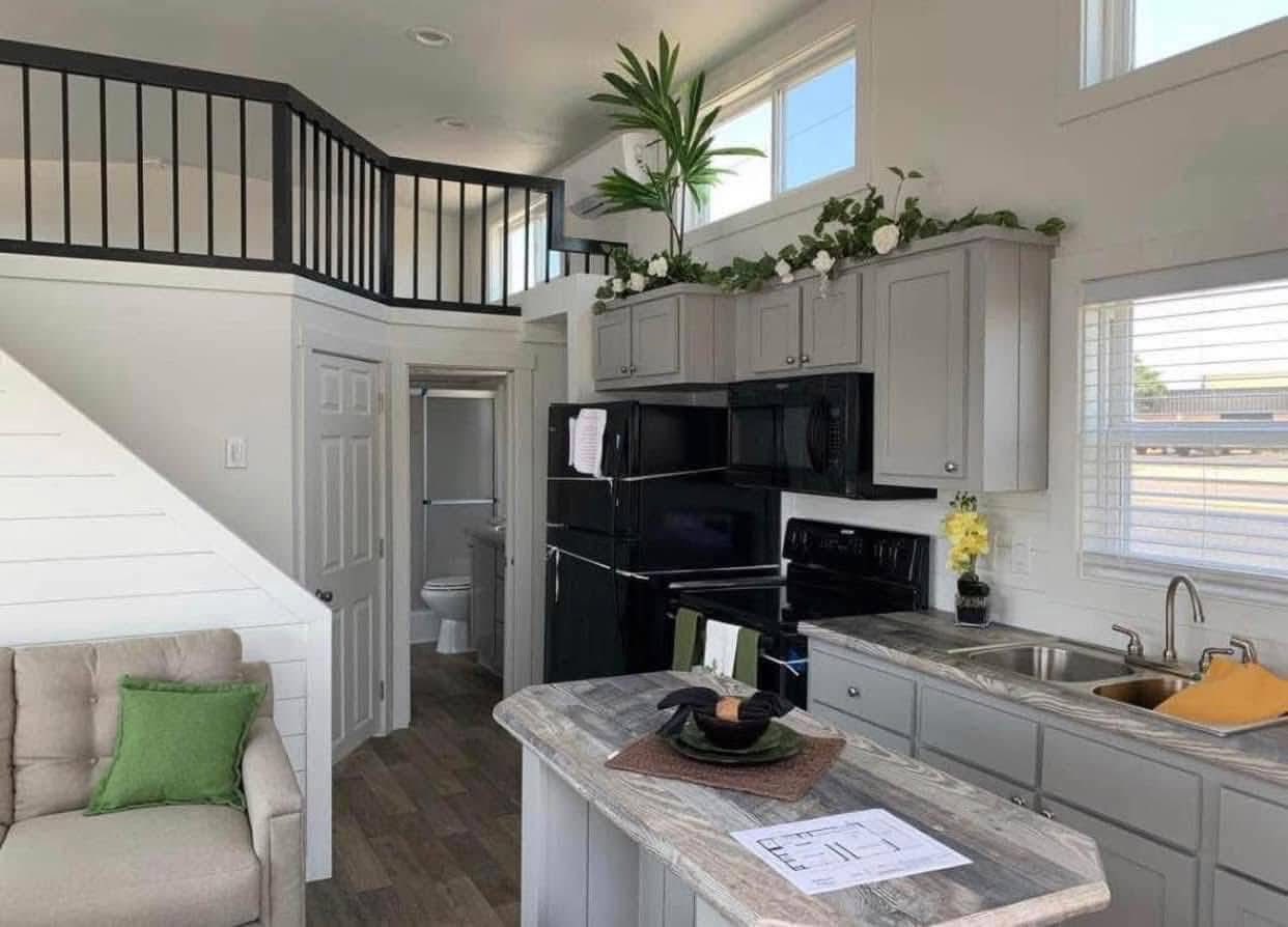 Modern tiny house interior with a loft. Gray kitchen cabinets adorned with plants. Cozy couch with green pillows. Bright and inviting atmosphere.