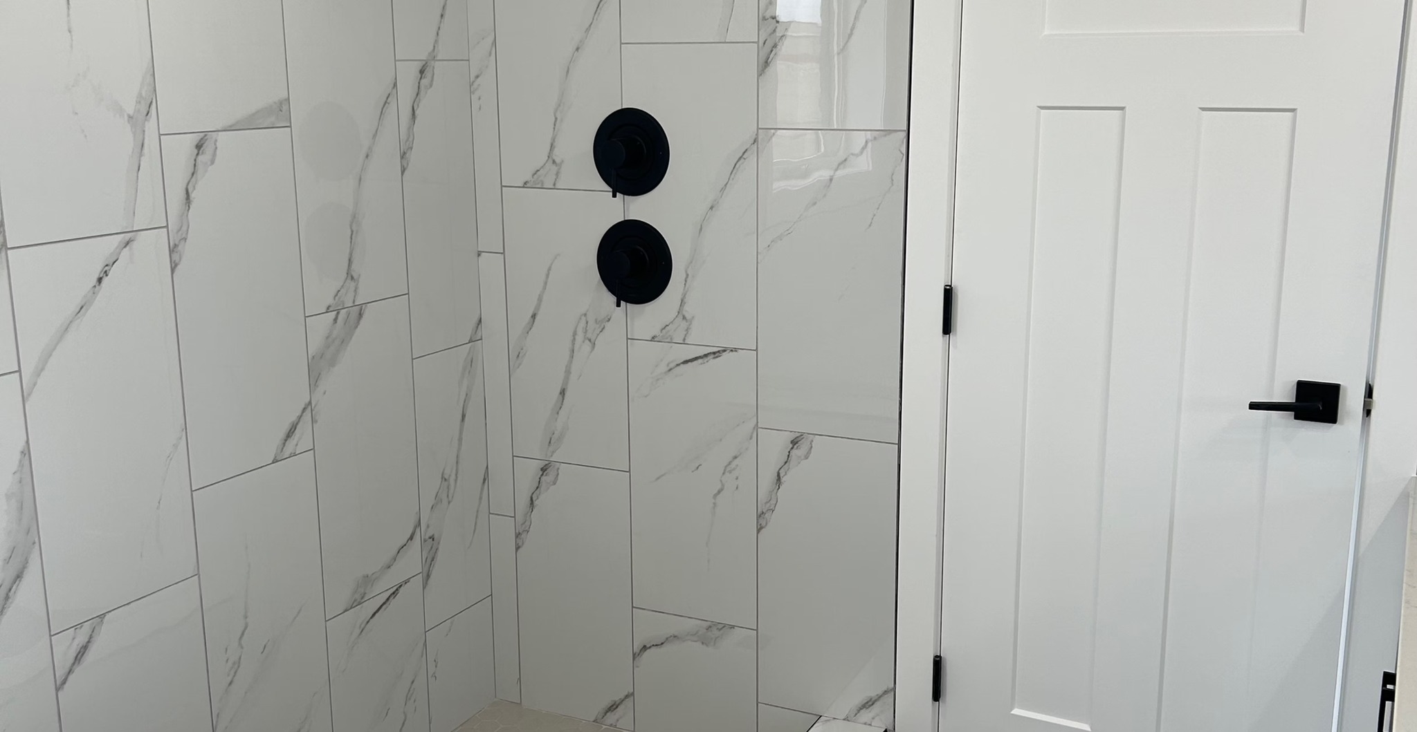 Sleek bathroom with glossy white marble tiles featuring gray veins, black matte handles, and a white wooden door. Modern and minimalistic atmosphere.