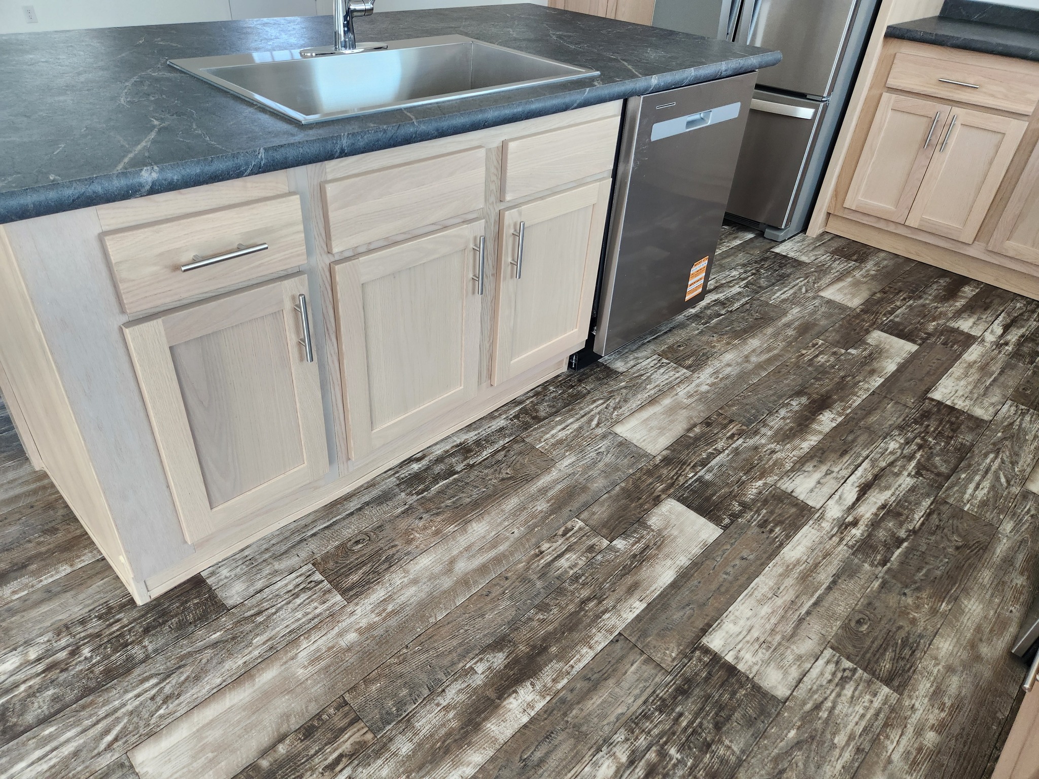 Modern kitchen interior with rustic wood-patterned flooring and light wood cabinetry. Features a black countertop, sink, and stainless steel dishwasher.