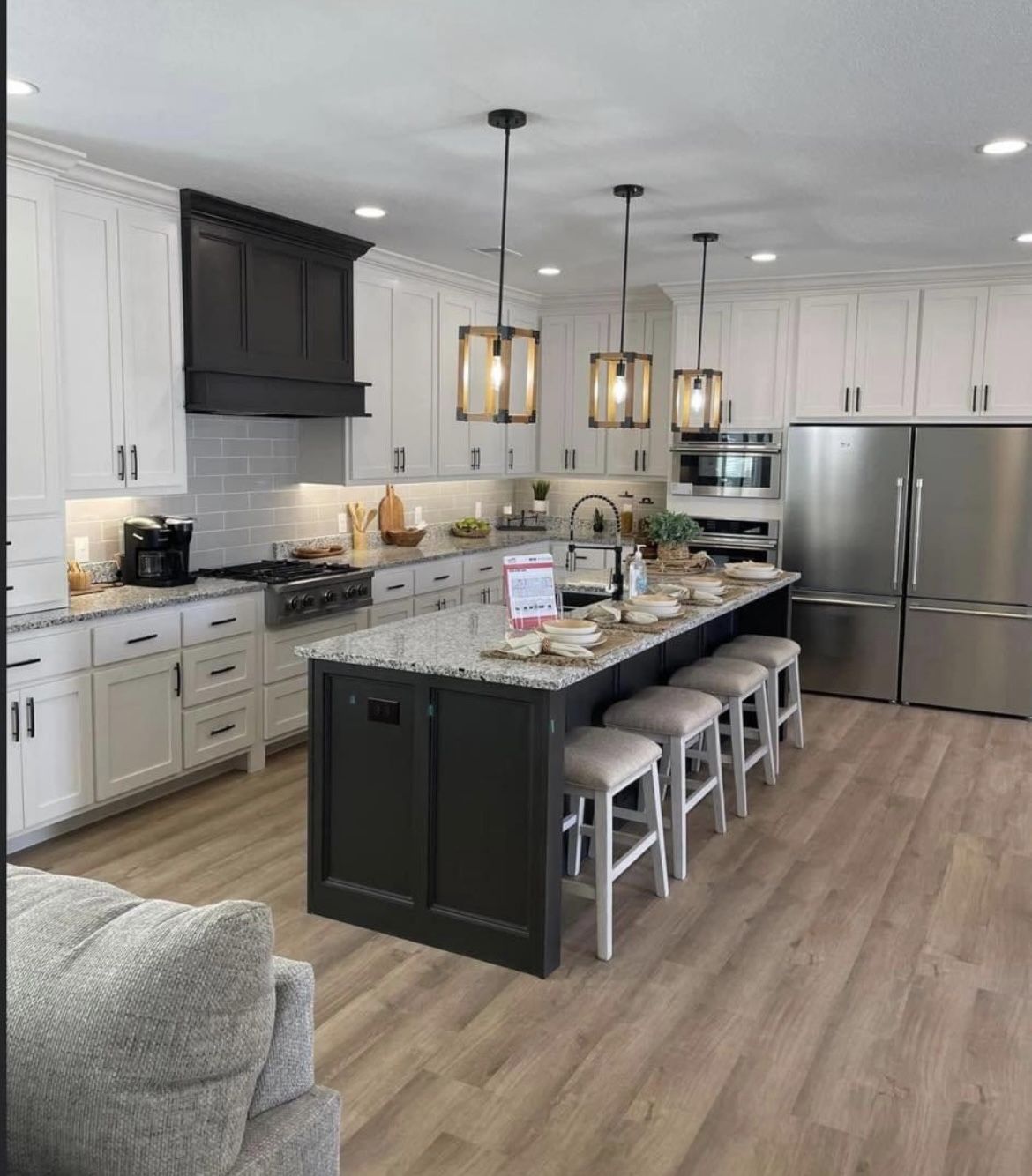 Modern kitchen with a large island, granite countertops, barstools, pendant lighting, stainless steel appliances, white cabinets, and wood flooring. Cozy and inviting.