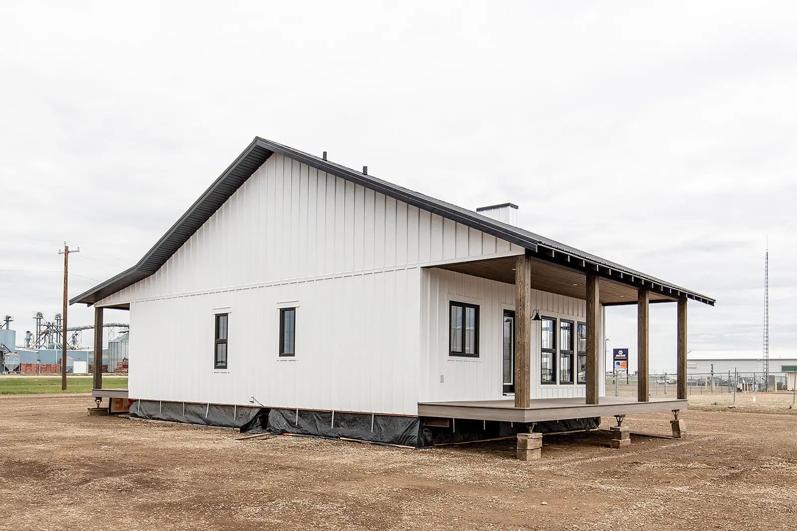 A modern, single-story house with white siding and a dark metal roof sits on a dirt plot. The house features a wraparound porch with wooden pillars, highlighting its contemporary design against a cloudy sky.