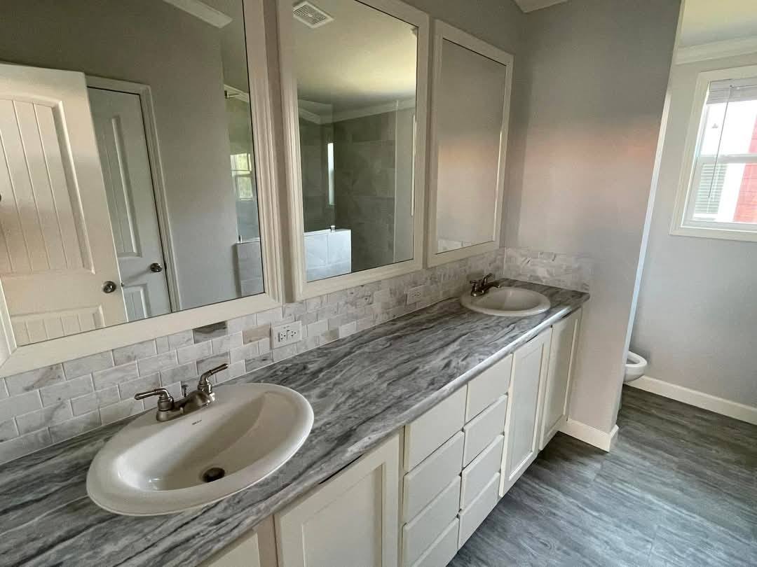 A modern bathroom with a sleek, gray marble countertop and dual sinks. Above are three mirrors, and bright natural light streams in from a window.