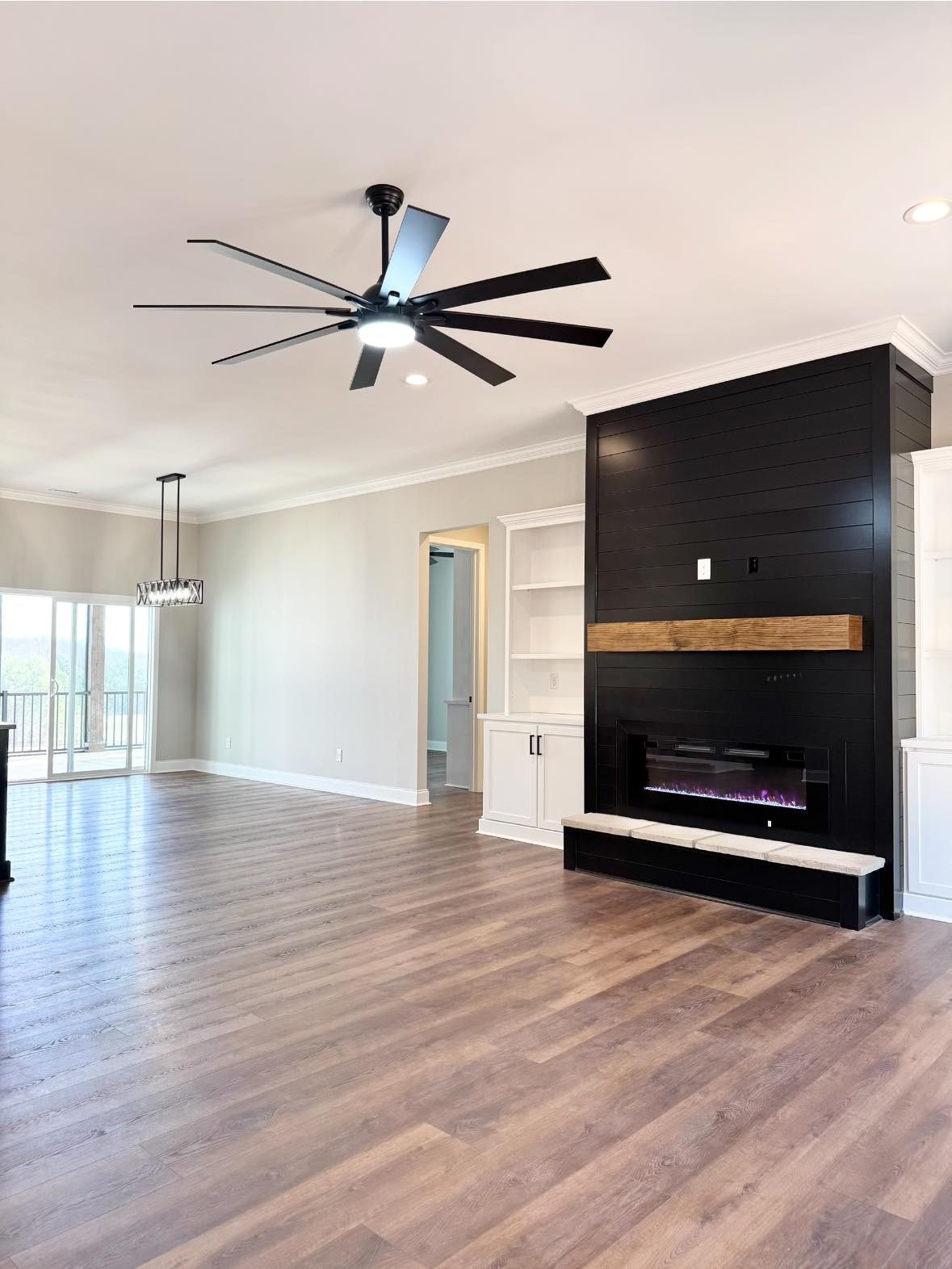 Spacious living room with wood floors, featuring a black shiplap fireplace, large ceiling fan, and modern chandelier. Bright and airy with natural light.