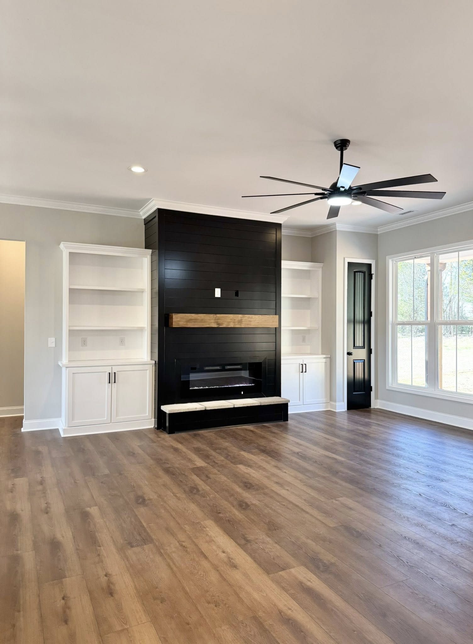 Spacious living room with wood flooring, black fireplace wall with a wooden mantel, surrounded by white cabinets. Ceiling fan and large windows create an airy feel.