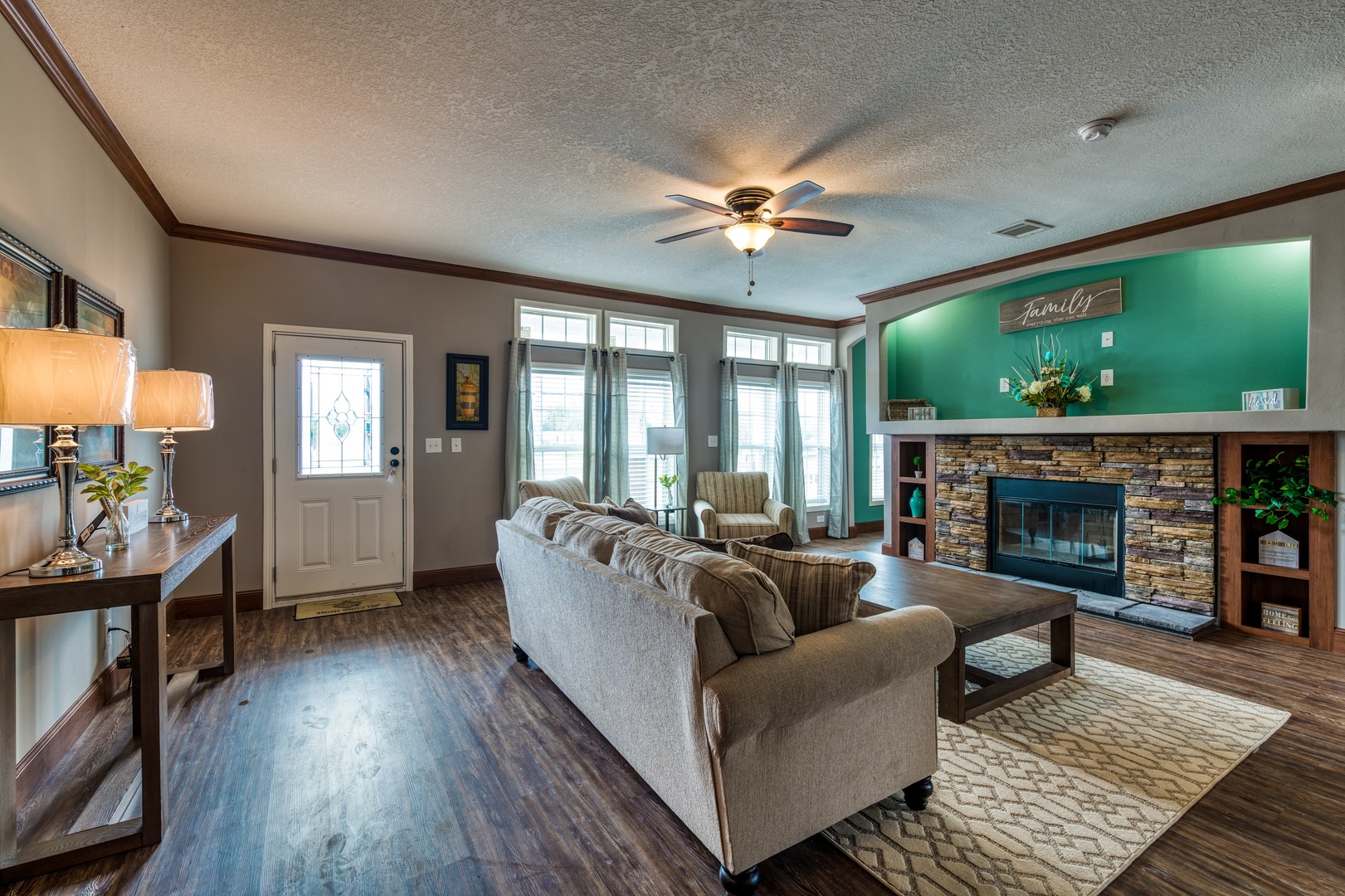 Cozy living room with wooden floors, beige sofa, and a stone fireplace. Ceiling fan, green accent wall, and soft lighting create a warm, inviting atmosphere.