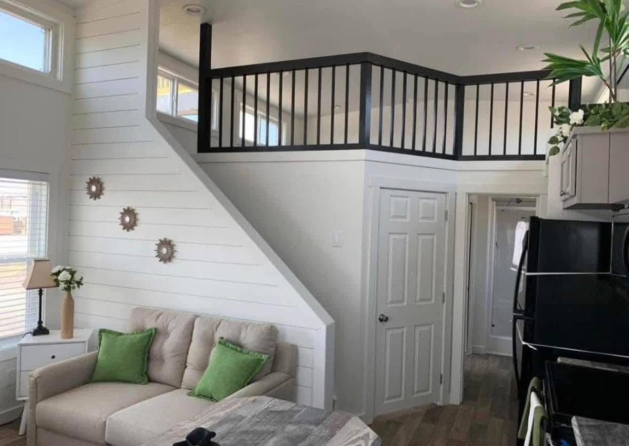 Cozy tiny house interior with a loft. Bright white walls, green-accented couch, and a modern black-railed staircase create an airy, inviting atmosphere.