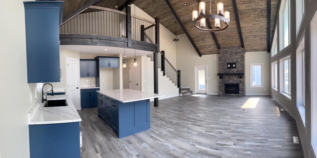 Spacious living area with high wooden ceilings and large windows. Features include a stone fireplace, modern blue kitchen cabinets, and a loft with railing. The room is airy, with a contemporary feel and natural lighting.