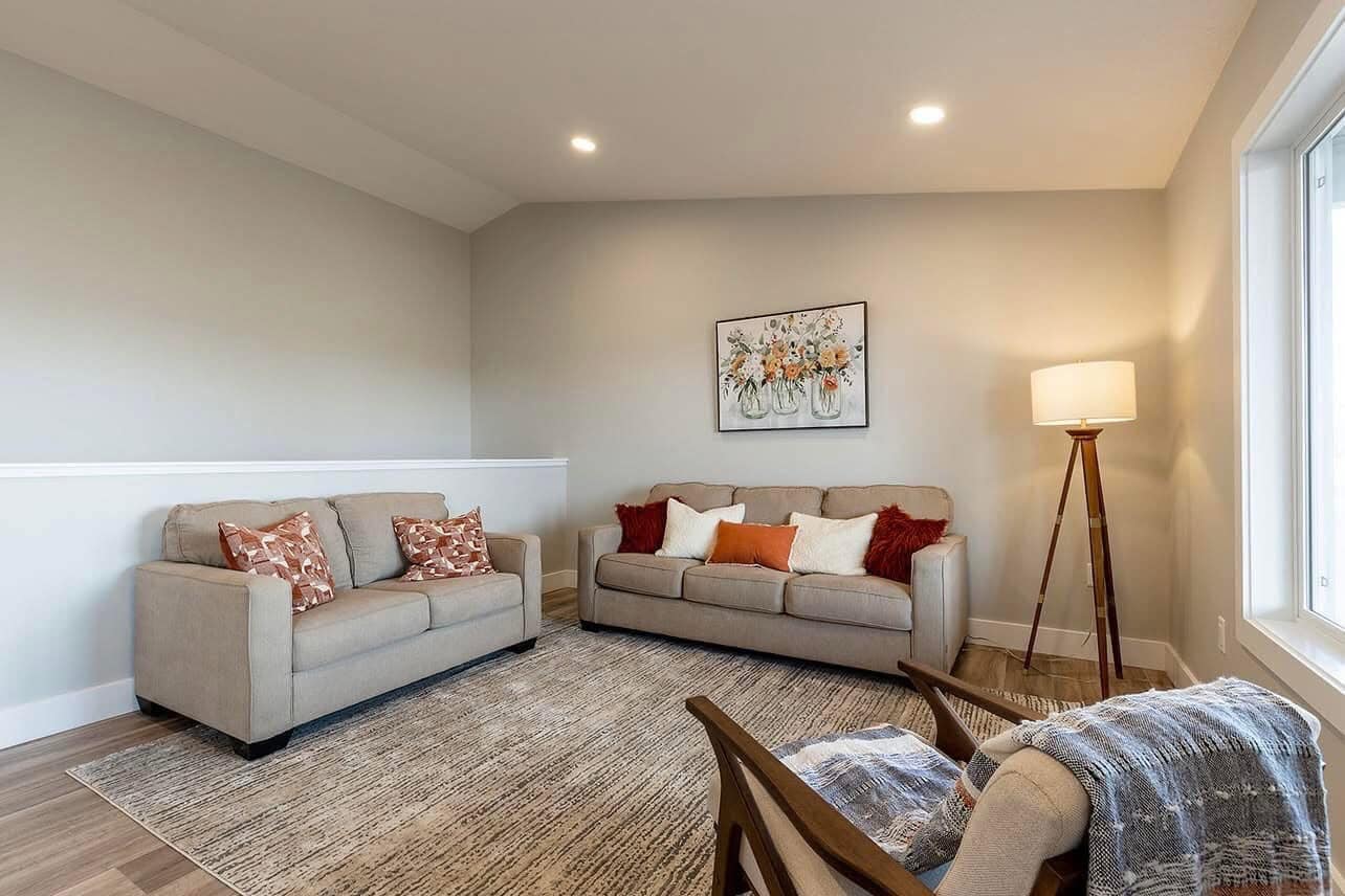 A cozy living room with beige walls features a gray sofa and loveseat adorned with patterned and solid pillows. A floral painting hangs above the sofa. A floor lamp emits warm light, and a soft rug covers the wooden floor. A wooden chair with a blanket faces the seating area, creating a welcoming atmosphere.