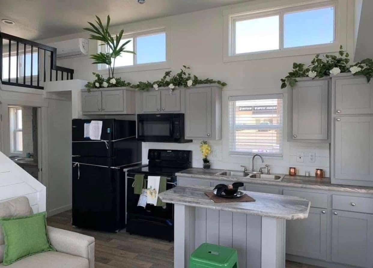 Modern kitchen with gray cabinets, black appliances, and a marble-top island. Green and white plants decorate above cabinets. Bright, airy ambiance.