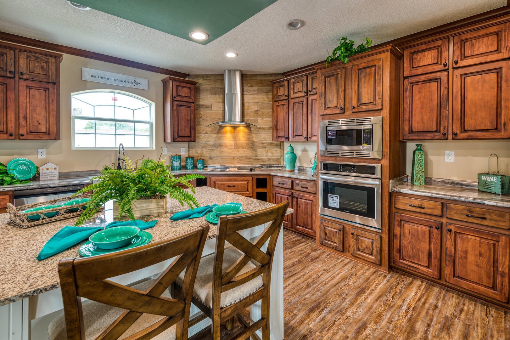 Warm kitchen with rich wooden cabinets, teal accents, and stone backsplash. Features a central island with green tableware, evoking a cozy, inviting feel.