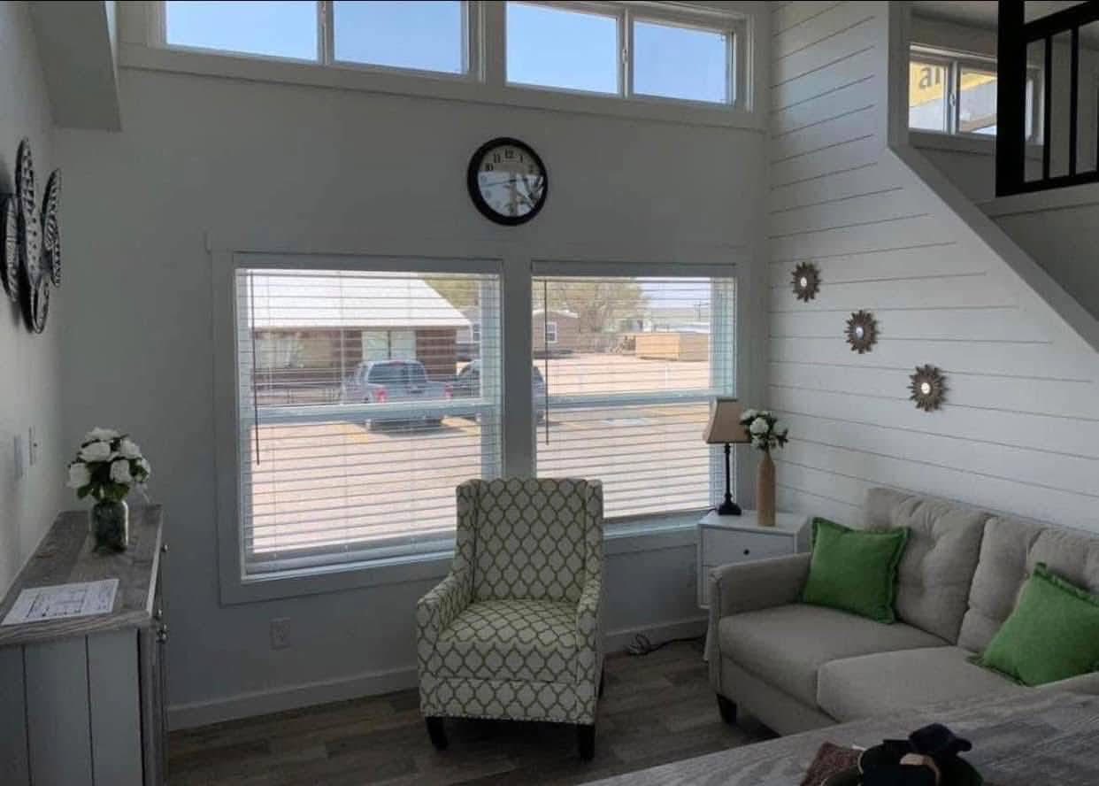Cozy living room with a large window, patterned green chair, beige sofa with green cushions, white shiplap walls, and decorative flowers. Bright and inviting.