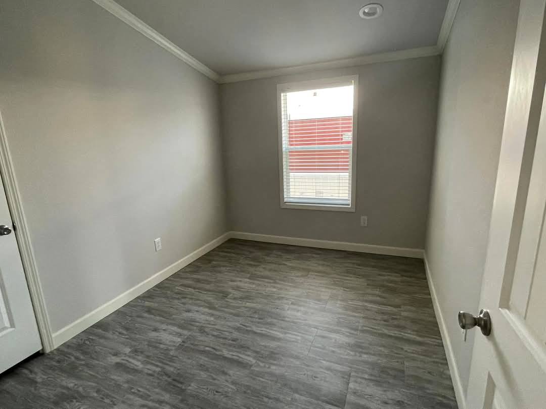 Small, empty room with gray walls and wood-like flooring. A single window lets in light, and a white door is partially open, creating a neutral, minimalist vibe.