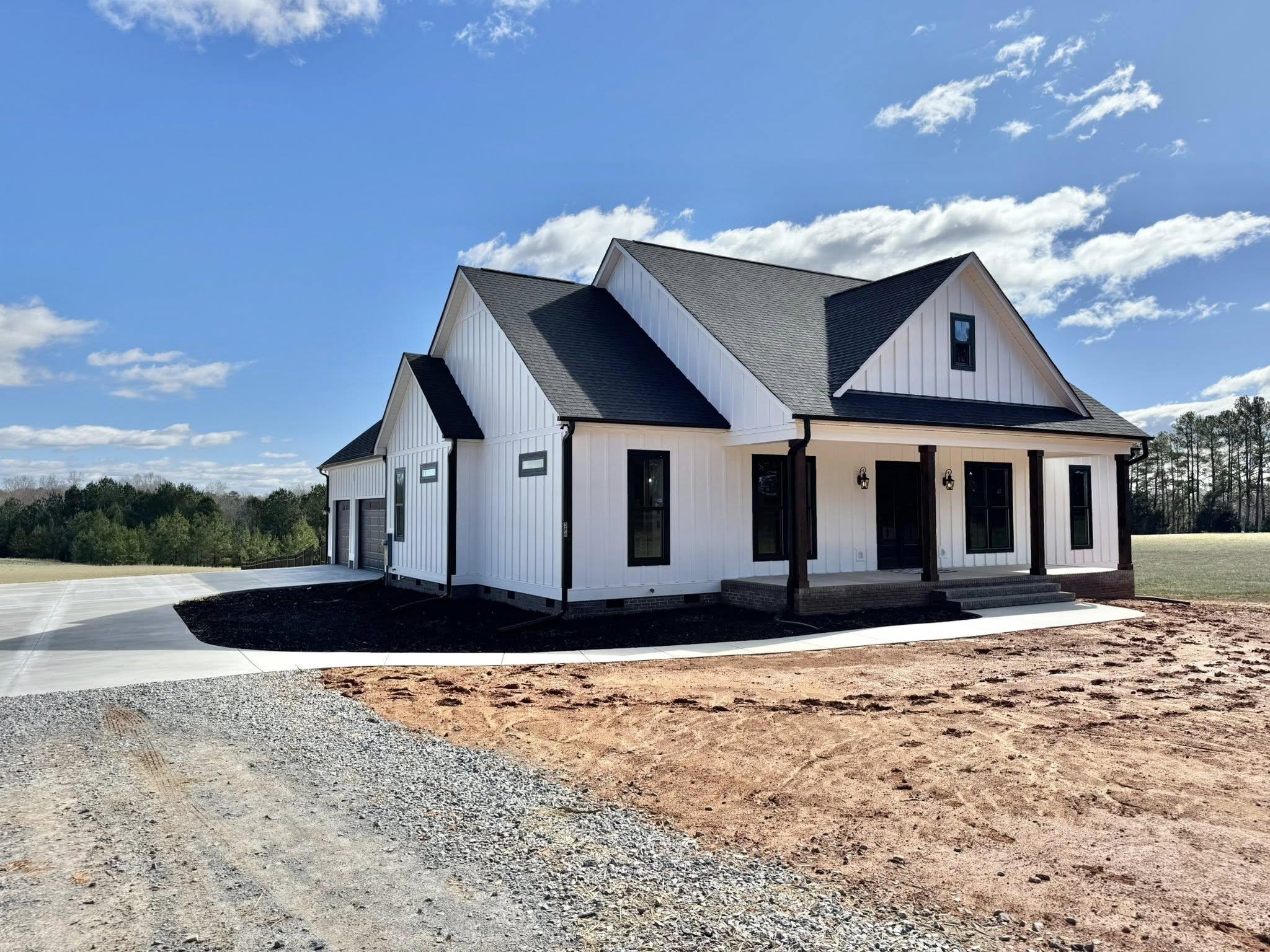 A modern white farmhouse with black trim and a covered porch sits under a blue sky. The surrounding area has a gravel driveway and open fields.
