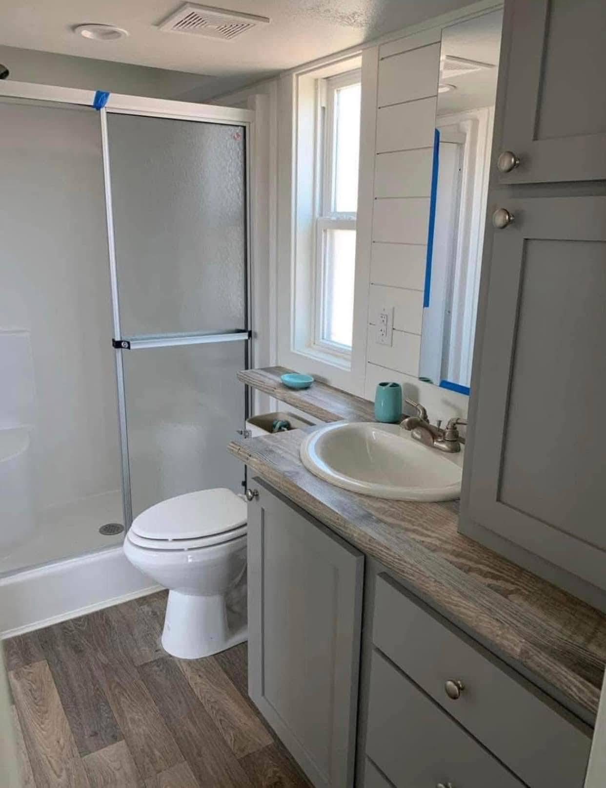 Compact, modern bathroom with a glass shower, white toilet, gray cabinets, and wood countertop. A window brings in natural light, creating a cozy feel.