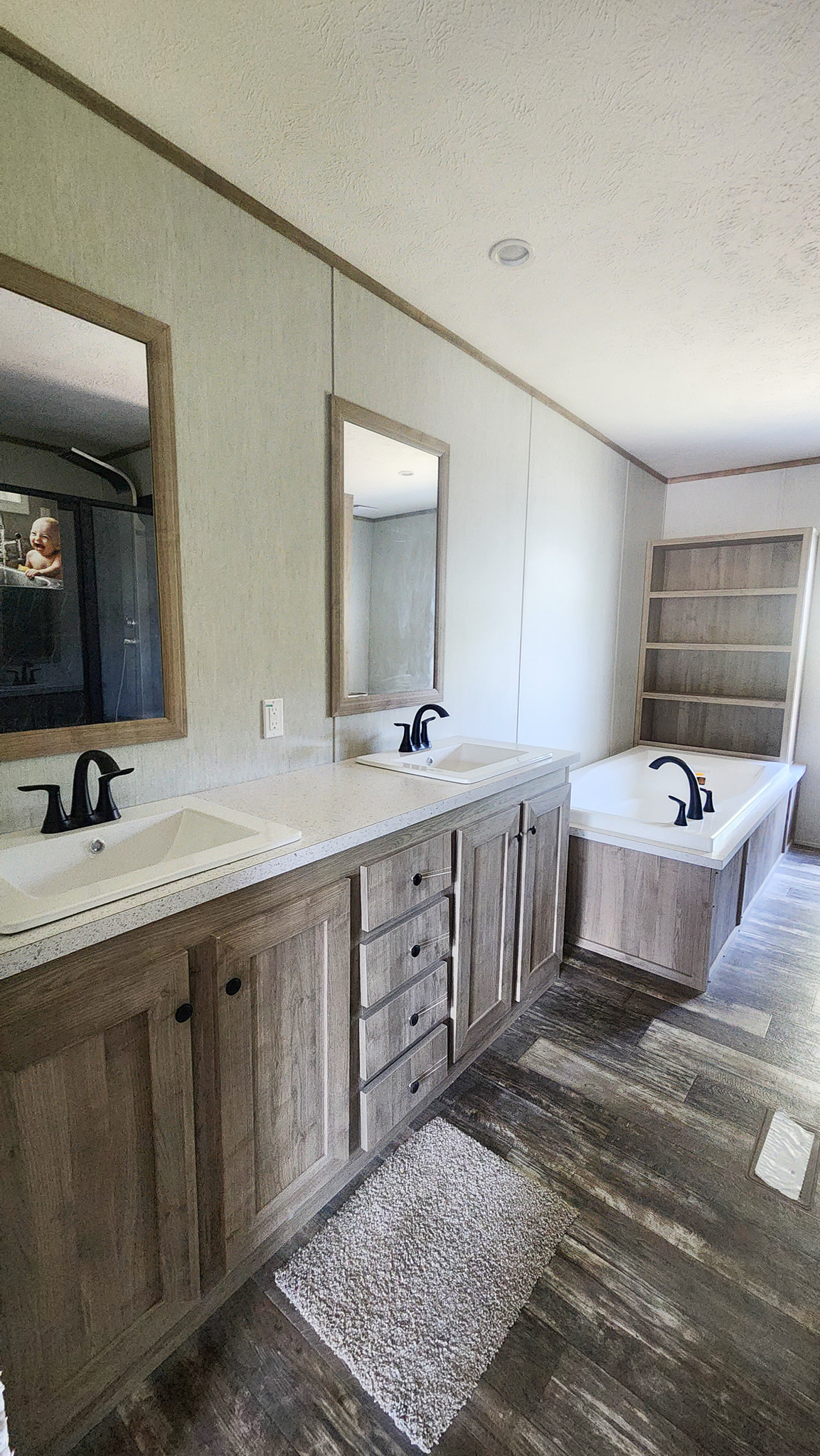 Modern bathroom with a rustic feel, featuring dual sinks with black faucets, a large mirror, soaking tub, wooden cabinets, and a plush rug on wood flooring.