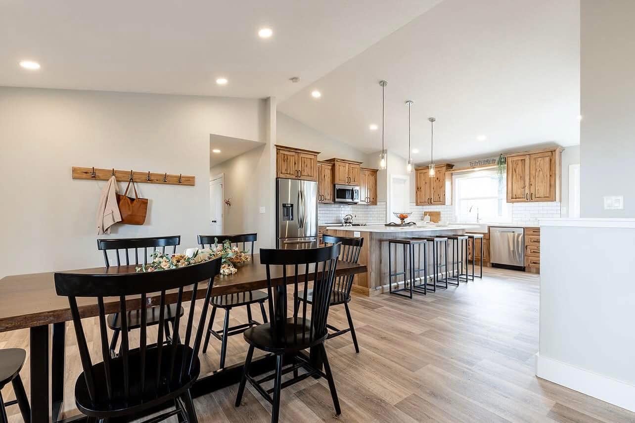 Open-concept kitchen and dining room with wooden furniture, black chairs, and light wood floors. Stainless steel appliances and pendant lights add a modern touch.