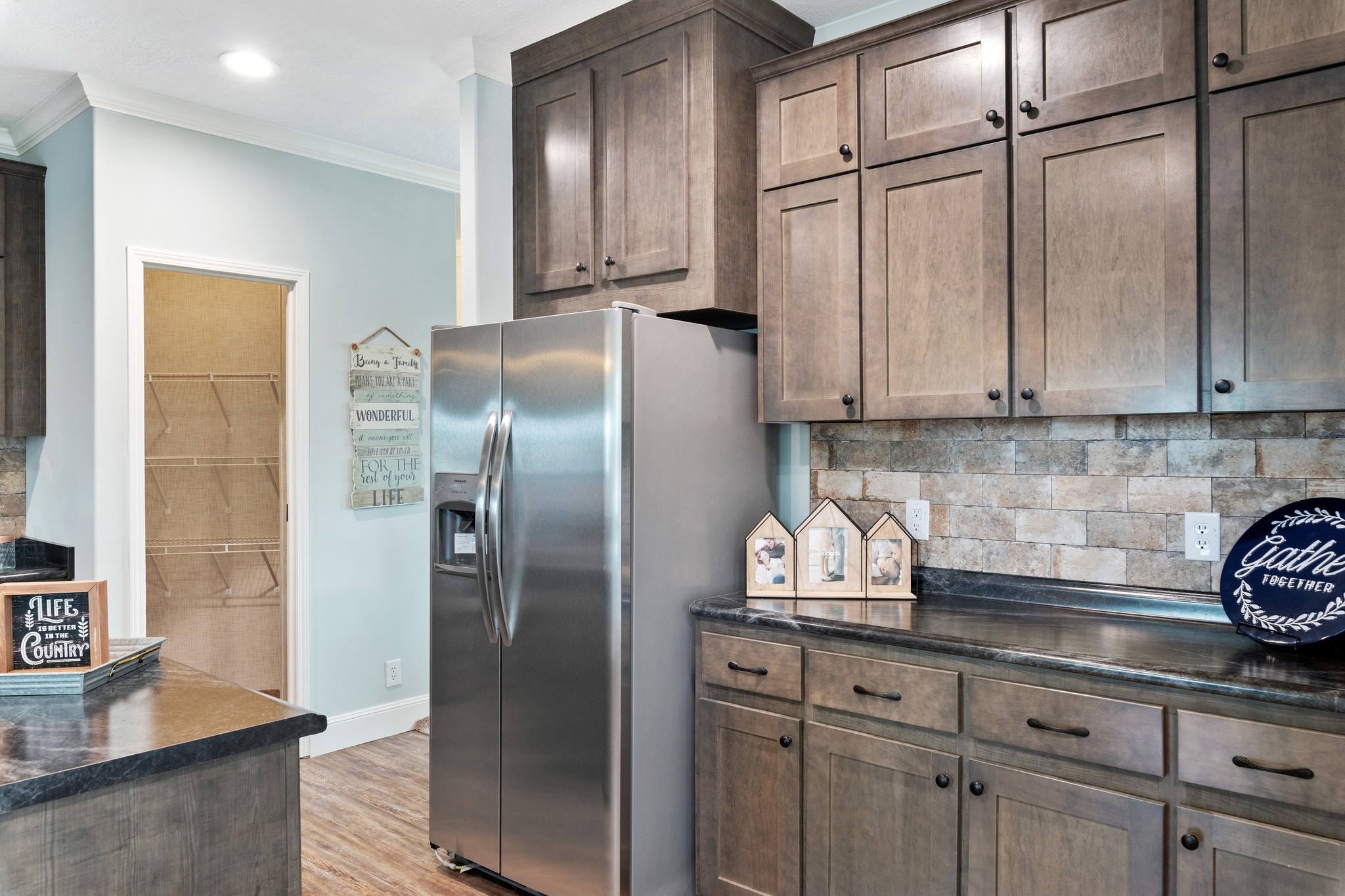 A cozy kitchen features light wood cabinets, a stainless steel fridge, and a stone backsplash. Décor includes small house ornaments and a rustic sign.