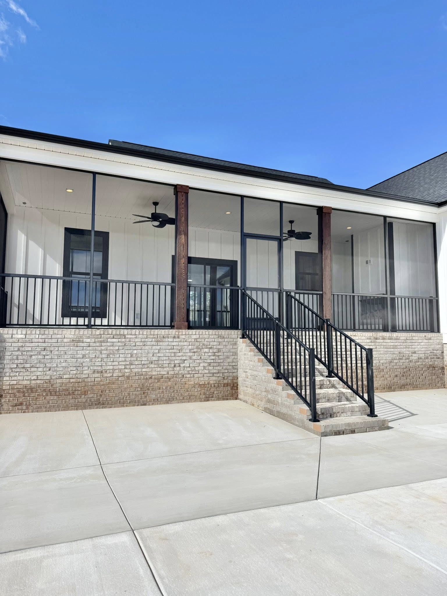 A modern house with white siding and a dark roof stands under a clear blue sky. It features a spacious porch with black railings and steps leading down to a patio.