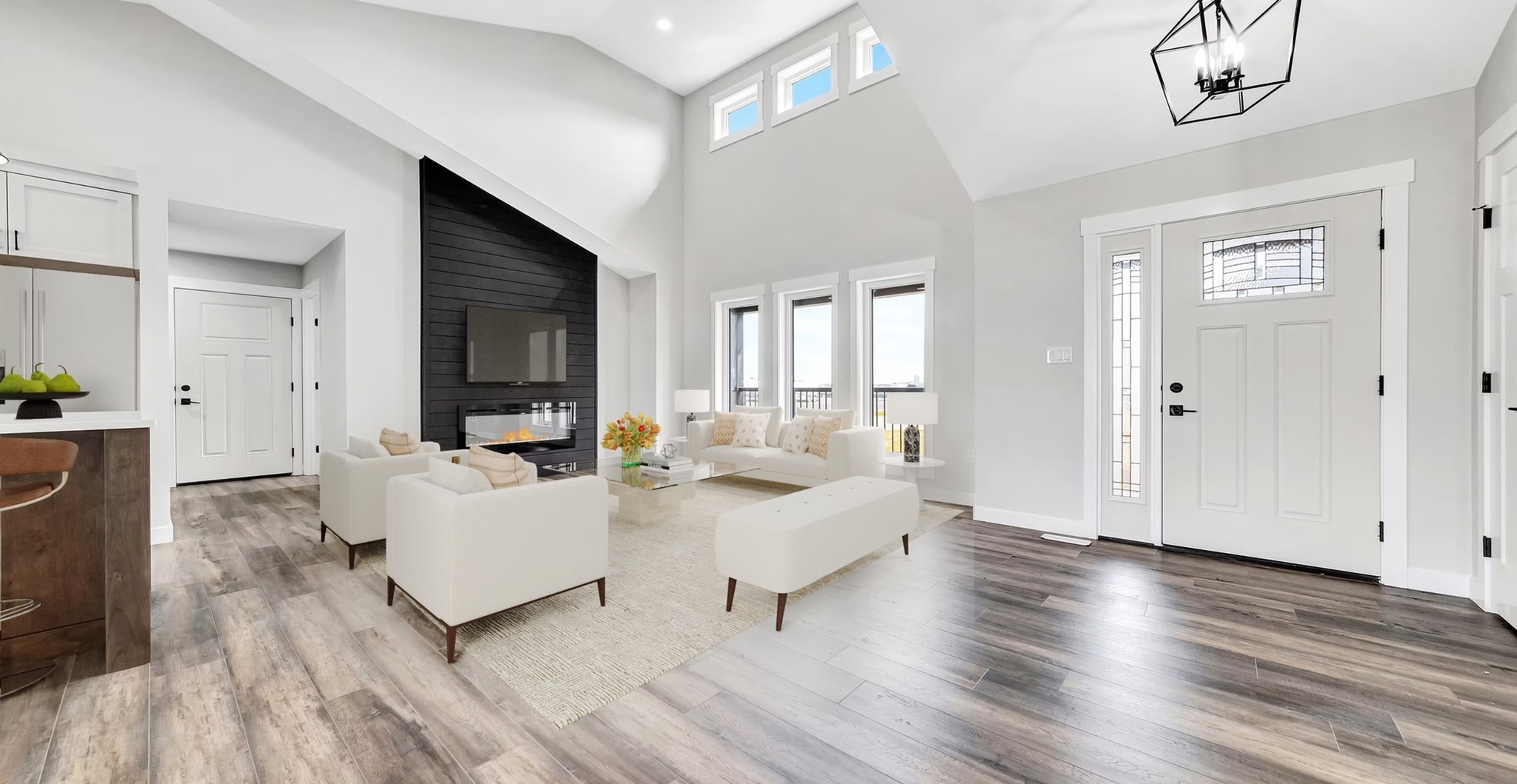 Bright, airy living room with high ceilings, wood floors, and modern decor. Features light furniture, a dark accent wall with a TV, and large windows.