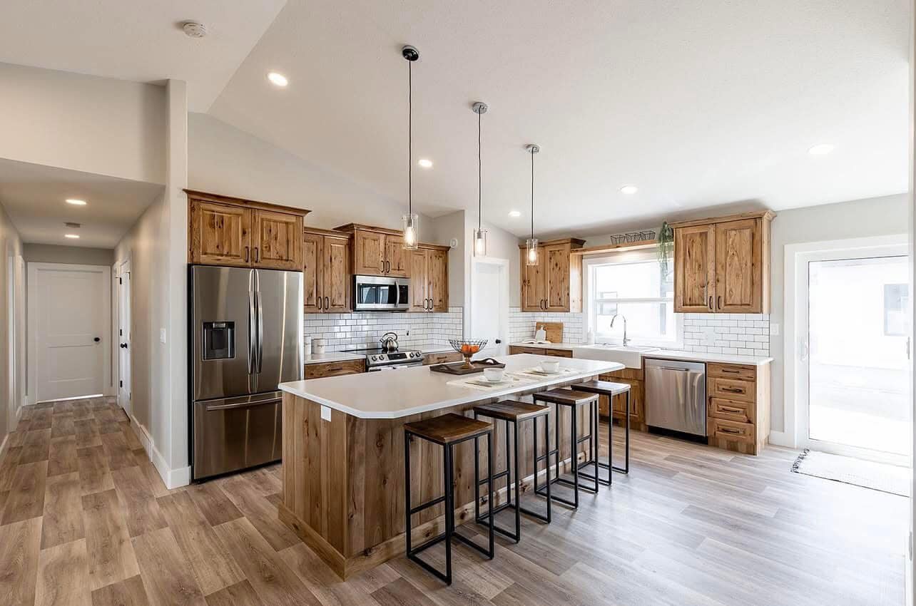 Spacious kitchen with rustic wooden cabinets, a stainless steel refrigerator, and a large island with bar stools. Bright lighting and neutral tones.