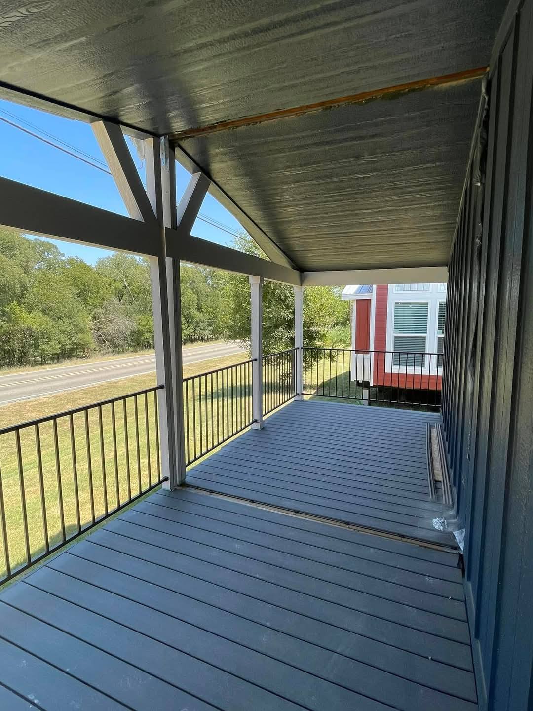 Covered wooden porch with black railings, overlooking a grassy yard and road. A nearby red and white house is partly visible. Clear, blue sky.