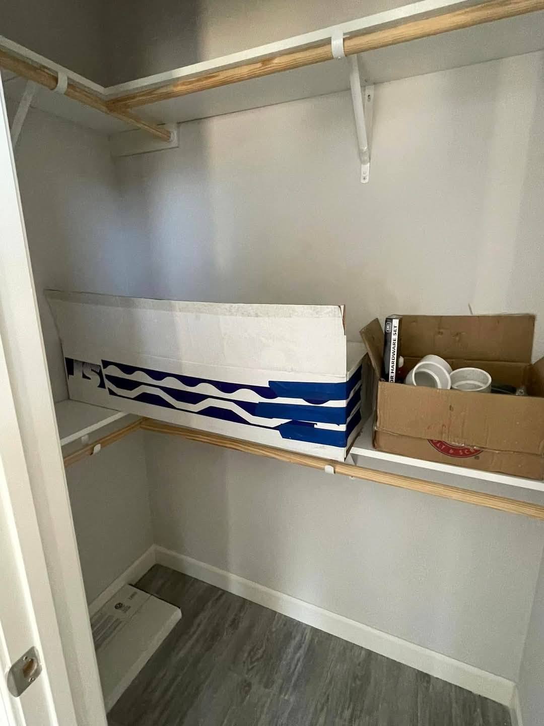 Small closet with wooden rods and gray walls. Contains two large boxes: one blue and white, one brown with dishes inside, resting on white shelves.