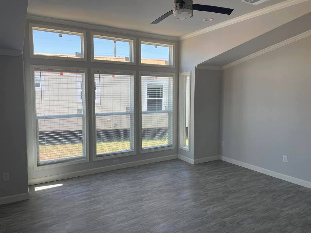 Spacious, modern room with large windows letting in natural light, light gray walls, and wood-look flooring. Ceiling fan adds a contemporary touch.