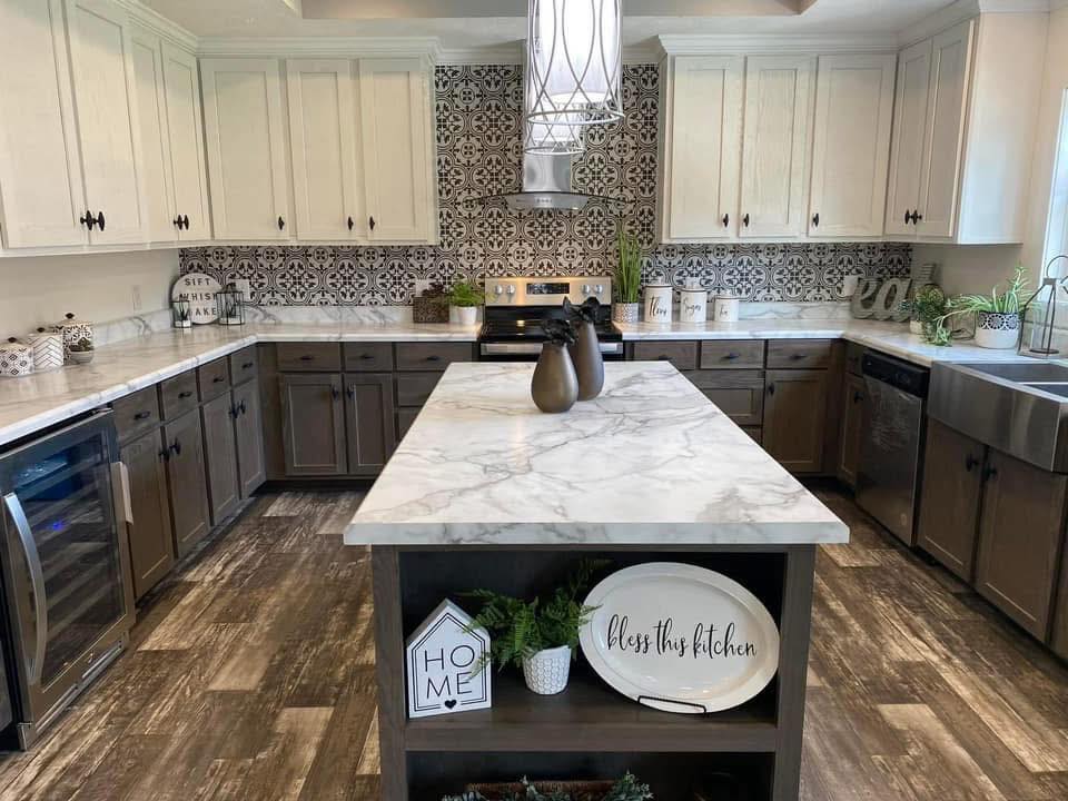 Modern kitchen featuring a marble island, patterned backsplash, rustic cabinets, and decorative signs. Warm lighting and greenery create a cozy, welcoming feel.