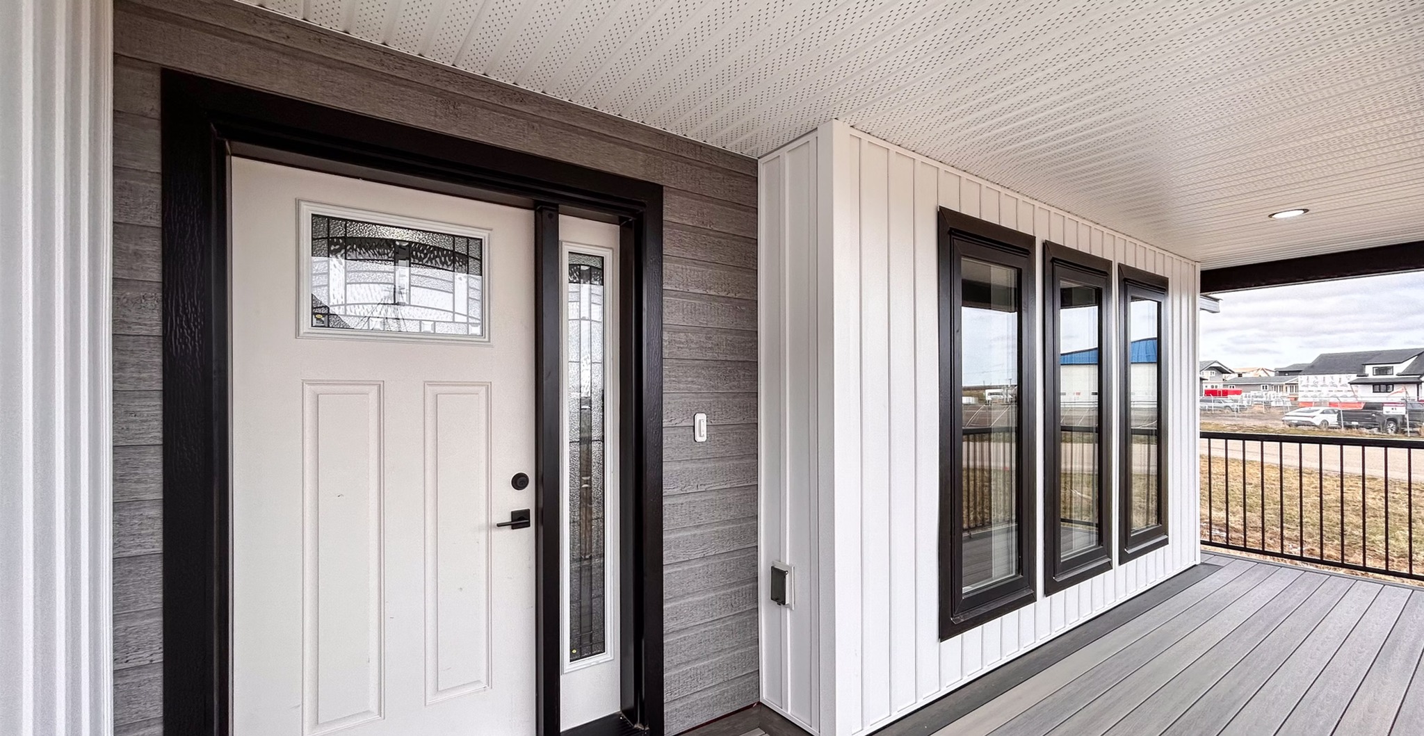 Modern porch with a white door, black trim, and adjacent vertical windows. Gray paneling and a wooden deck create a sleek, inviting entryway.