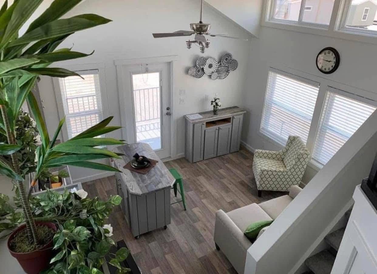 Bright, modern living room with wood floors, a large potted plant, cozy armchair, small grey table, wall clock, and window letting in natural light.