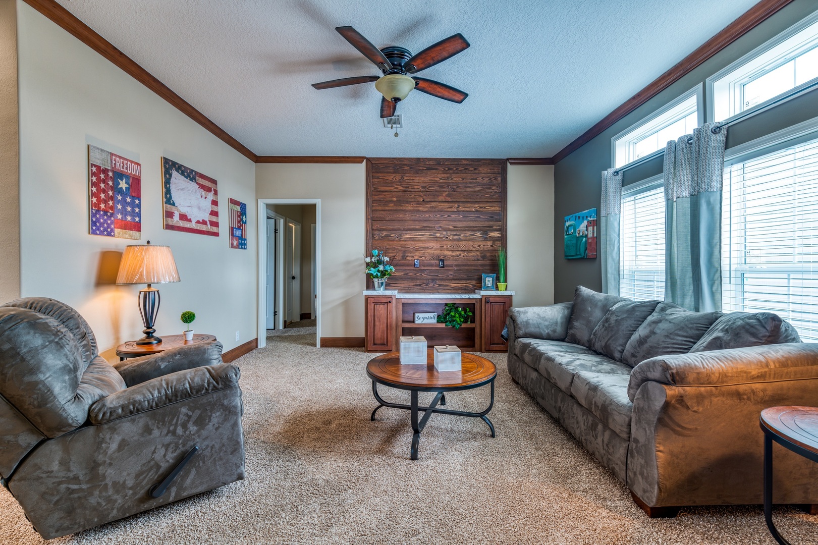 Cozy living room with beige walls, a brown sectional sofa, and a recliner. A round wooden coffee table sits on a plush carpet. Large windows with curtains let in natural light. A ceiling fan hangs above, and patriotic-themed art decorates the walls.