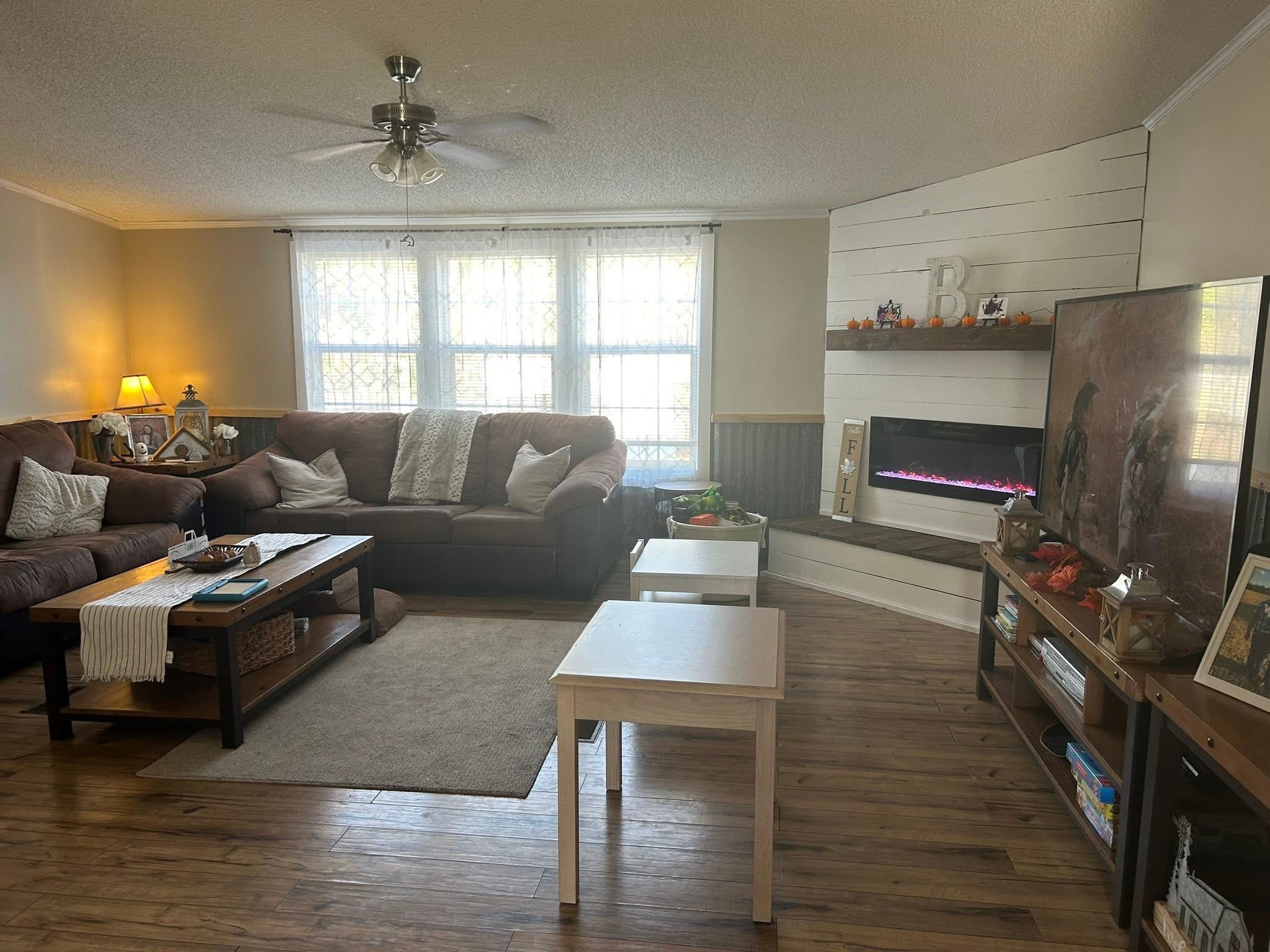 Cozy living room with brown sofas, cushioned armchair, and coffee table on a beige rug. Electric fireplace on a white paneled wall, large window with sheer curtains, TV, and wooden floors; warm, inviting atmosphere.