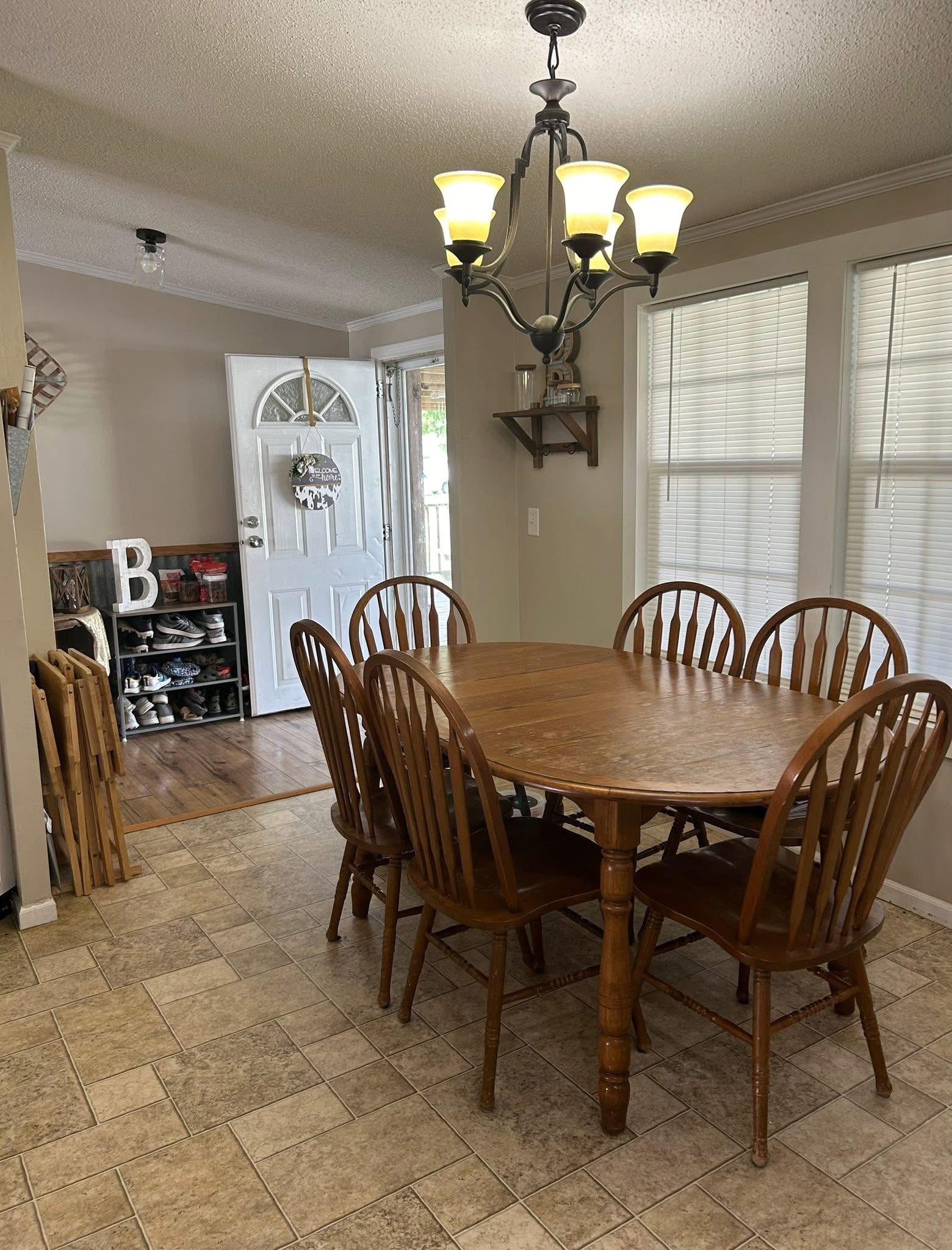 Dining room with a wooden table and six matching chairs under a lit chandelier. An open front door reveals a shoe rack and welcome sign, creating a warm, inviting atmosphere.