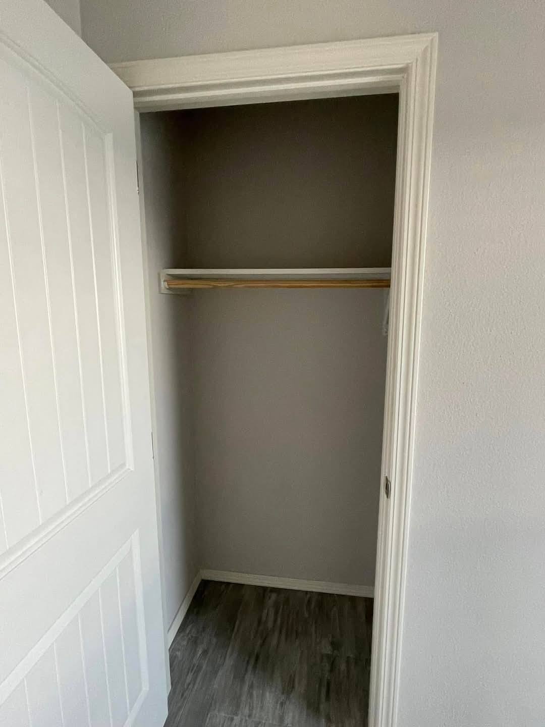 Empty closet with open white door, displaying a wooden hanging rod. The walls are light gray, and the floor has a dark wood pattern, creating a minimalist feel.