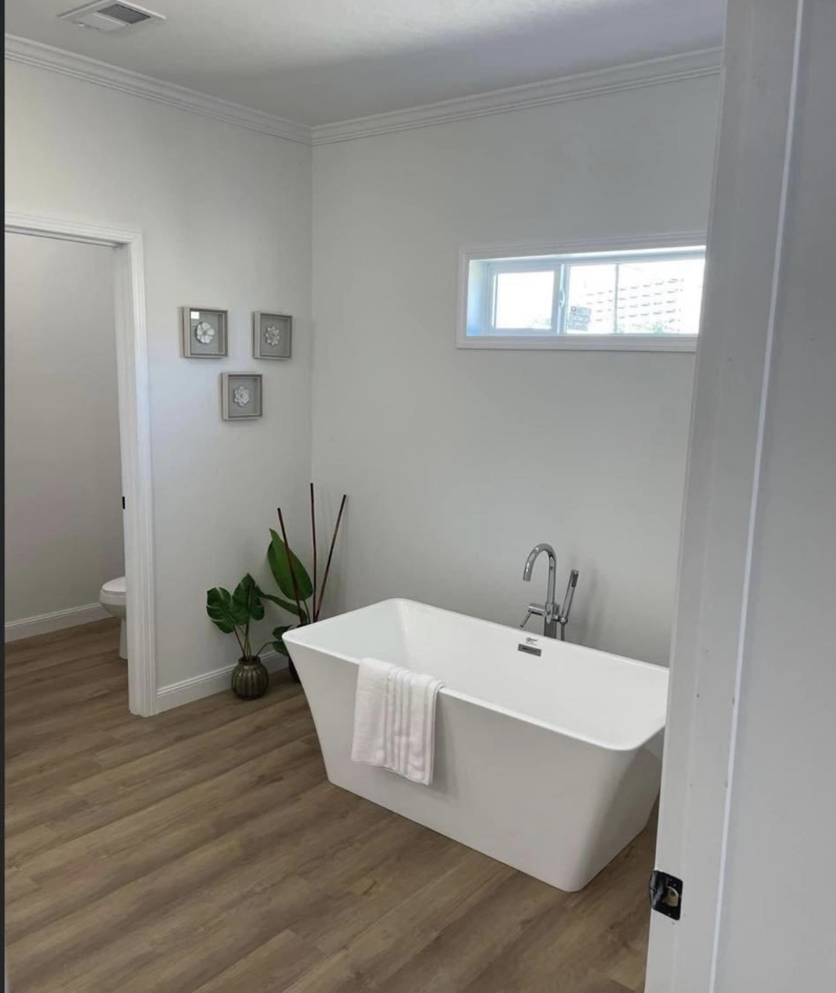 Minimalist bathroom with a freestanding white bathtub on wood flooring. A towel drapes over the bathtub. A small window and wall art add elegance.