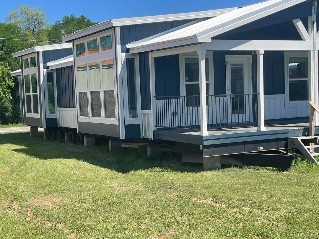 Modern blue and white modular home with large windows sits elevated on a grassy lawn. It has a small porch, evoking a welcoming and cozy feel.