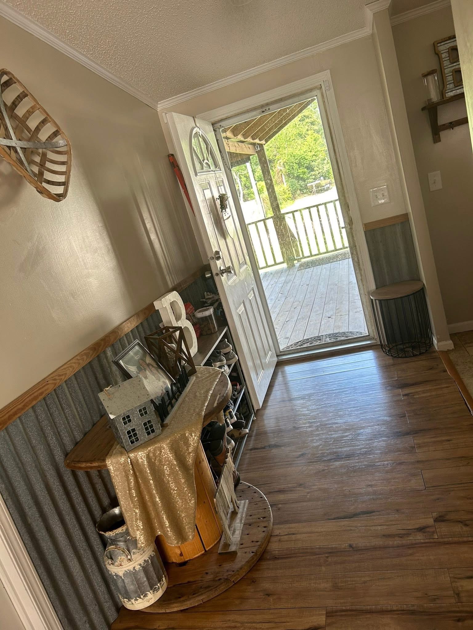 Sunny hallway leading to an open door, revealing a porch and greenery. Inside, decor includes a small shelf with a gold cloth, mini house, and rustic accents.