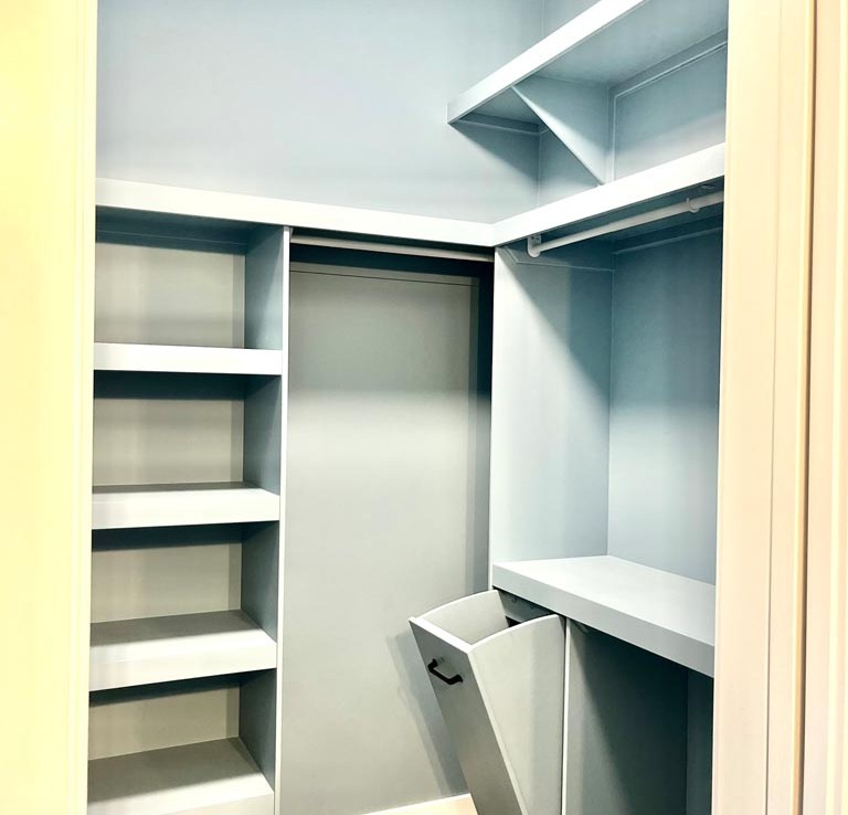 A small, organized closet with light blue walls, featuring shelves on the left, a hanging rod on the right, and a tilt-out hamper beneath. Modern and tidy.
