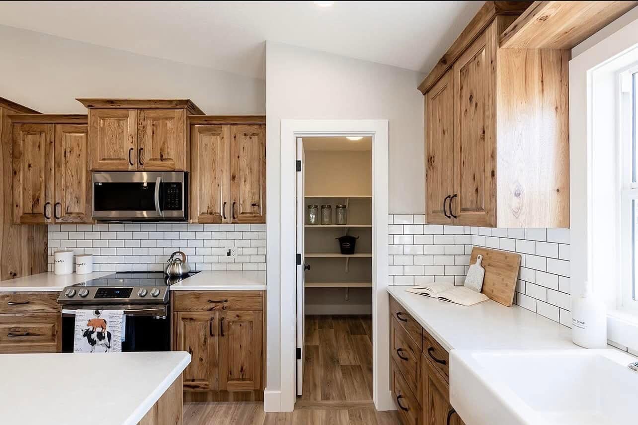 Rustic kitchen with natural wood cabinets, white countertops, and subway tile backsplash. A pantry is visible through an open door, creating a welcoming and warm atmosphere.