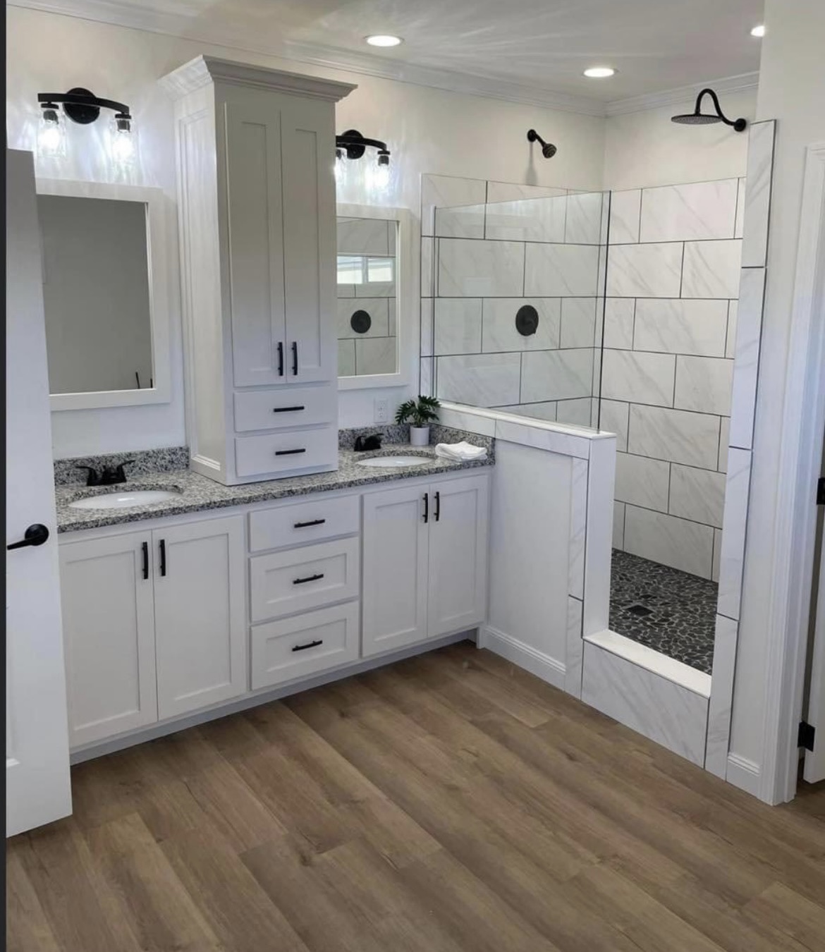 Modern bathroom with white cabinets, granite countertops, and wood flooring. There's a walk-in shower with large tiles and sleek black fixtures.