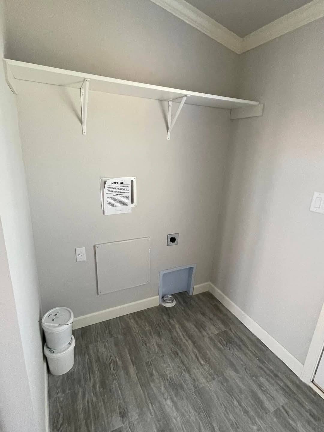 Empty laundry room with light gray walls, a white shelf, and wood-look flooring. A notice is posted on the wall near stacked plastic containers.