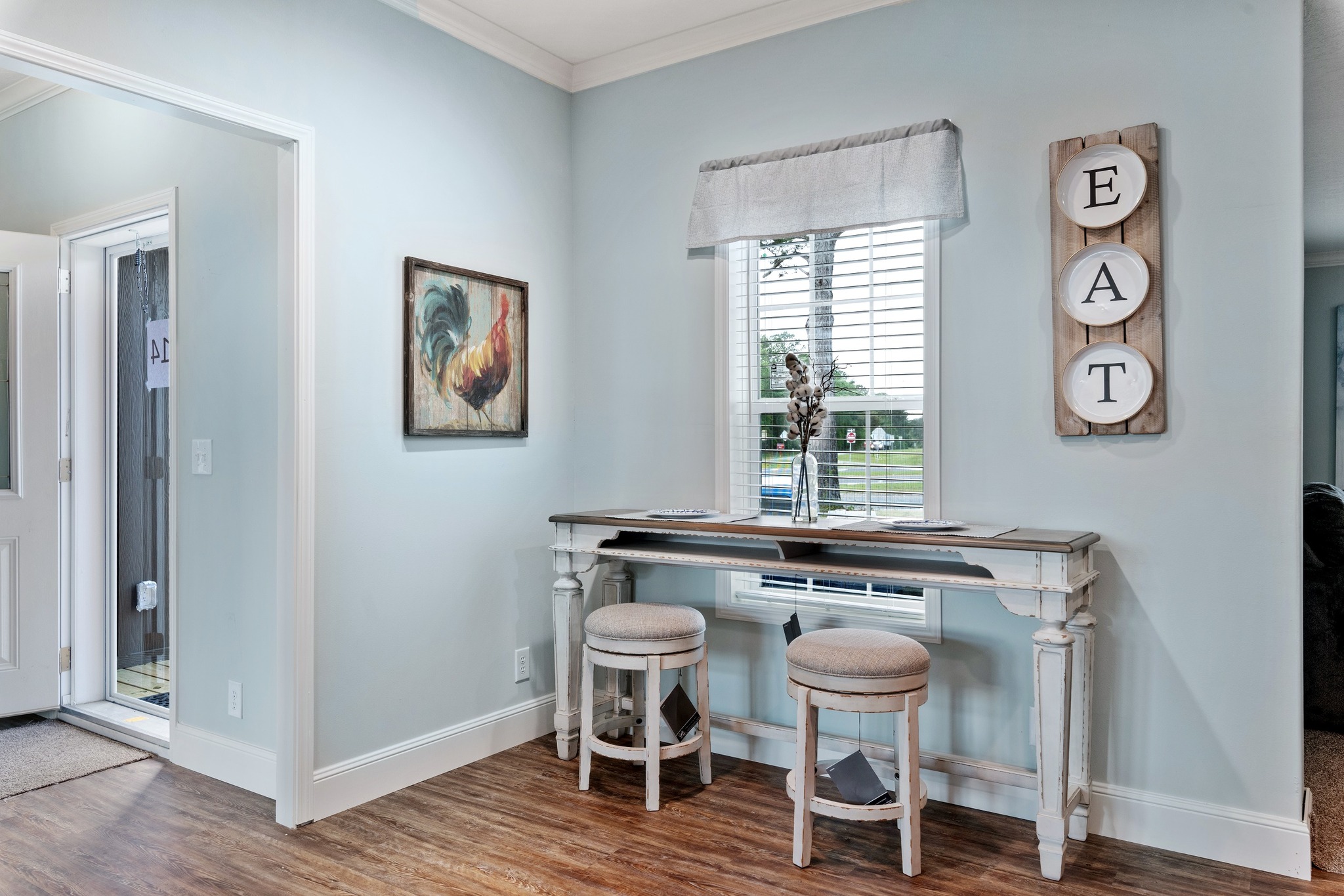 A cozy kitchen nook with light blue walls features a rustic wooden table and two cushioned stools under a window. A rooster painting and "EAT" wall decor add charm.