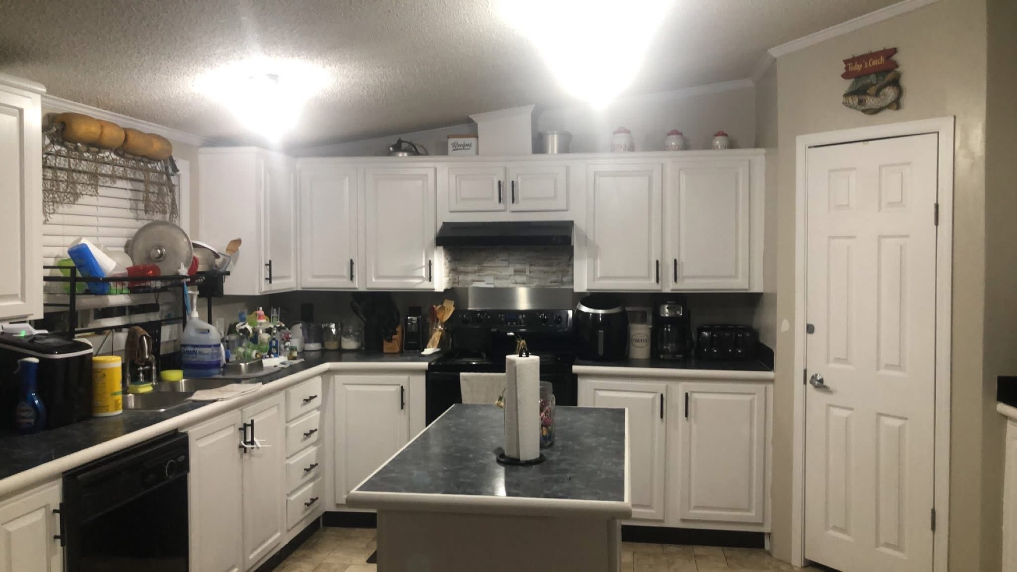 Modern kitchen with white cabinets and black countertops, centered island with paper towels, and various kitchen items. Bright lighting creates a clean, organized feel.