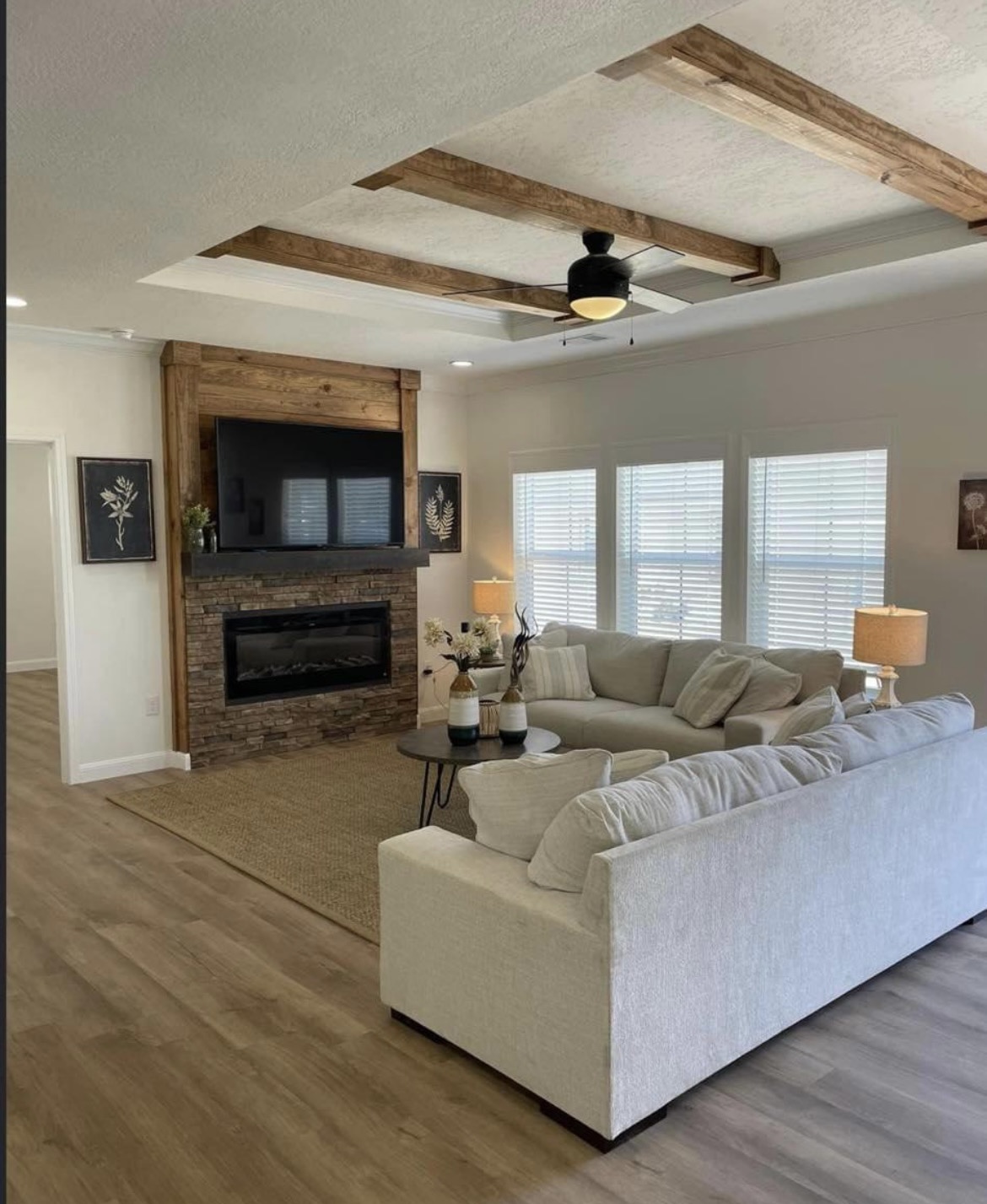 Cozy living room with a beige sectional sofa, stone fireplace, and TV. Exposed wooden beams on the ceiling and natural light create a warm atmosphere.