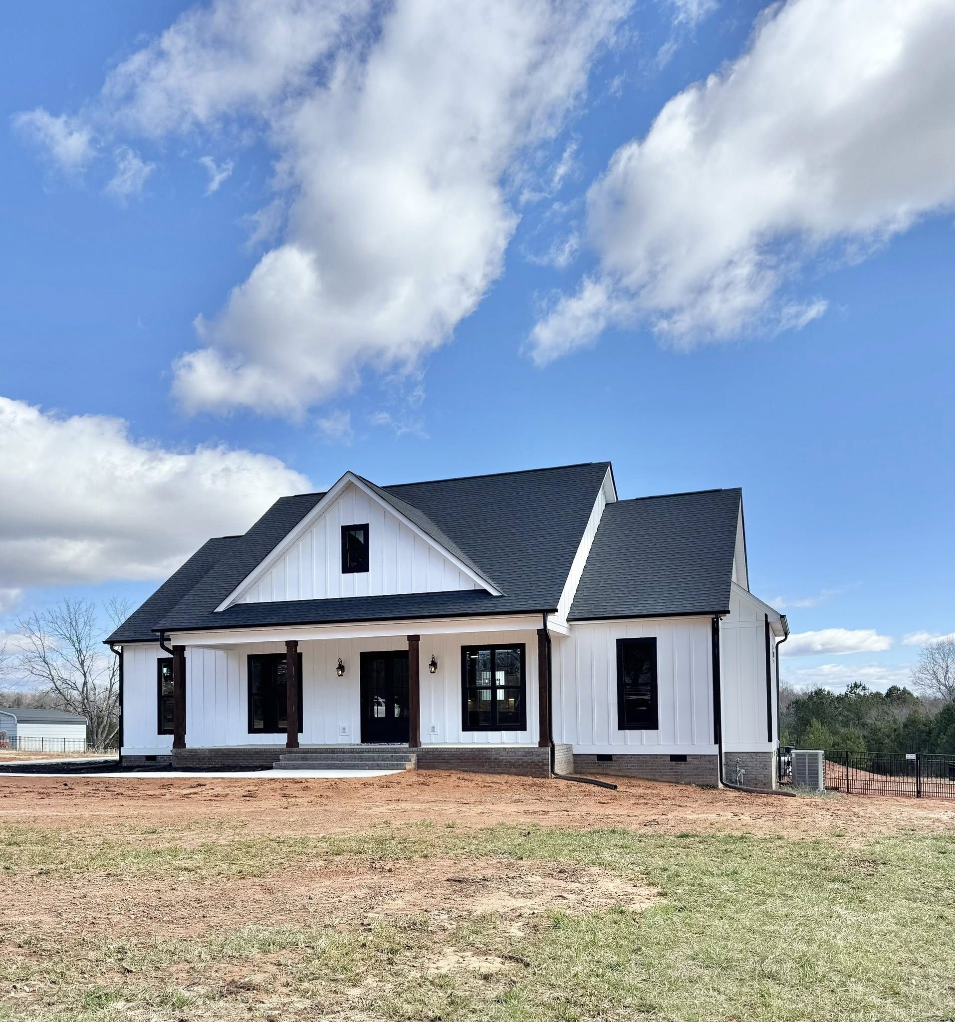 A modern farmhouse with white siding and a black roof sits under a blue sky with scattered clouds. The front porch features dark window frames.